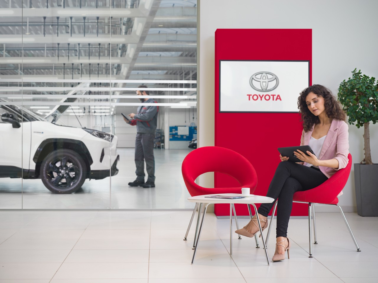 Žena sedí v čakárni servisu Toyota, pozerá sa na tablet a na televíznej obrazovke za ňou sa zobrazuje logo Toyota.